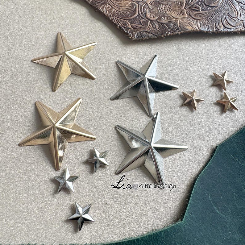 May include: Assortment of gold and silver star-shaped embellishments. The stars are various sizes and are arranged on a white surface. The text "Lia - simpledesign" is visible in the bottom right corner of the image.