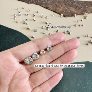 May include: A variety of copper-set glass rhinestone rivets in different sizes and colors. The rivets are displayed on a neutral background, with some held in a hand. The text "Copper-Set Glass Rhinestone Rivet" is visible.