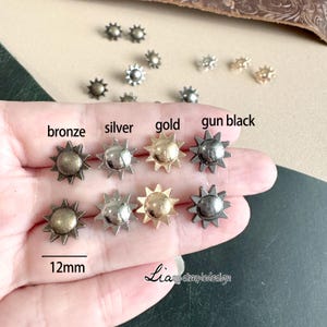 Puede incluir: Surtido de tachuelas metálicas en forma de estrella de 12 mm en acabados bronce, plata, oro y negro mate. Las tachuelas están dispuestas en una fila sobre una mano, con los nombres de los colores etiquetados debajo de cada tachuela.