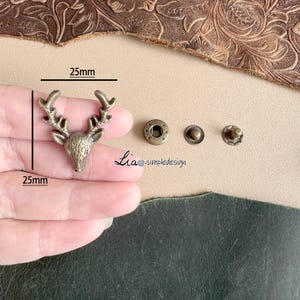 May include: A gold-toned metal deer head charm with a 25 mm measurement. The charm is shown on a hand with three additional metal snaps, each with a diameter of approximately 10 mm.