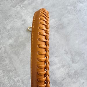 May include: Close-up of a tan leather handbag handle with a braided design. The handle is a warm, light brown colour and features a gold-coloured metal clasp. The stitching is visible along the edges.