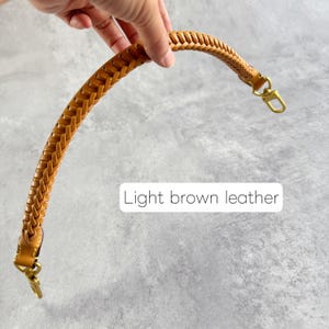 May include: A light brown leather bag handle with a plaited design. The handle features gold-coloured metal clasps at each end. The text "Light brown leather" is displayed in a white rectangle.