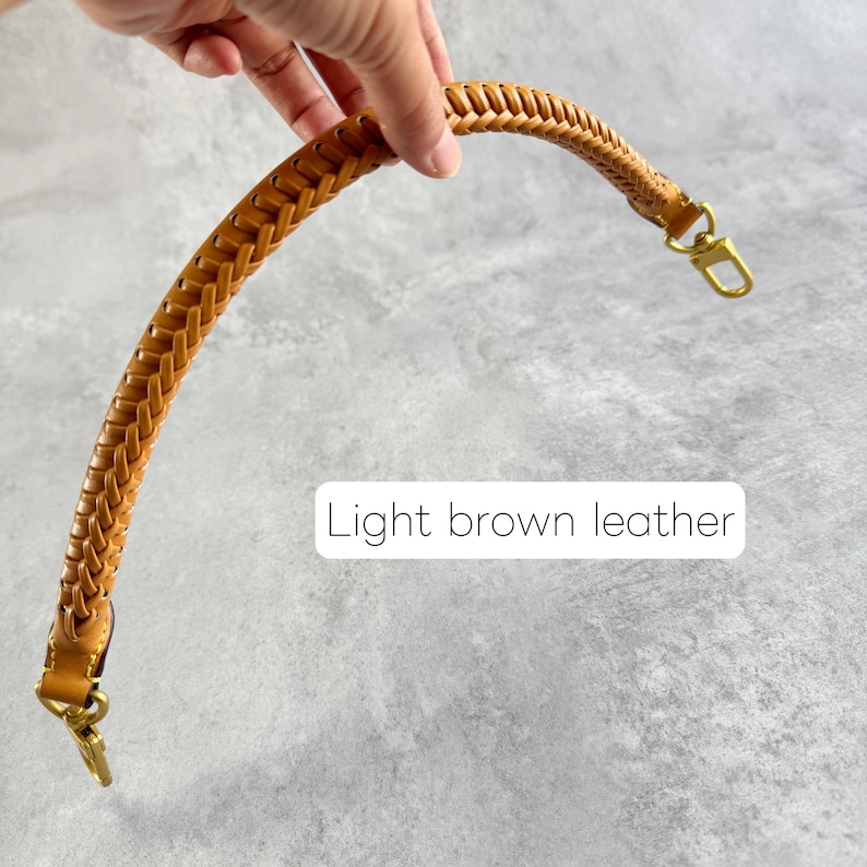 May include: A light brown leather bag handle with a plaited design. The handle features gold-coloured metal clasps at each end. The text "Light brown leather" is displayed in a white rectangle.