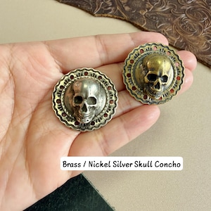 May include: Two round conchos featuring a central human skull design. One concho is finished in a metallic brass tone, while the other is finished in a nickel silver tone. Both pieces have a decorative scalloped edge with small circular cutouts.