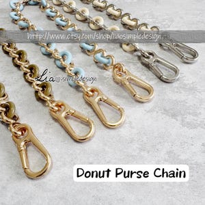 Può includere: Primo piano di diverse catene per borse con diversi design e stili di chiusura. Le catene presentano maglie dorate, argentate e verde oliva, alcune con accenti a forma di ciambella azzurra. Il testo "Donut Purse Chain" è visibile.