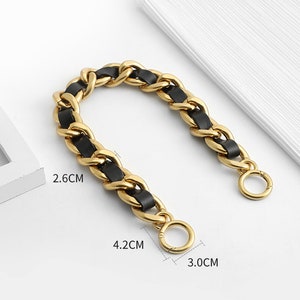 Peut inclure: Une cha&icirc;ne en m&eacute;tal dor&eacute; entrelac&eacute;e d'une bande en similicuir noir. La cha&icirc;ne comporte deux anneaux circulaires &agrave; ressort aux extr&eacute;mit&eacute;s. Les dimensions indiqu&eacute;es sont 2,6 cm, 4,2 cm et 3,0 cm, pr&eacute;cisant la taille des maillons et des anneaux.