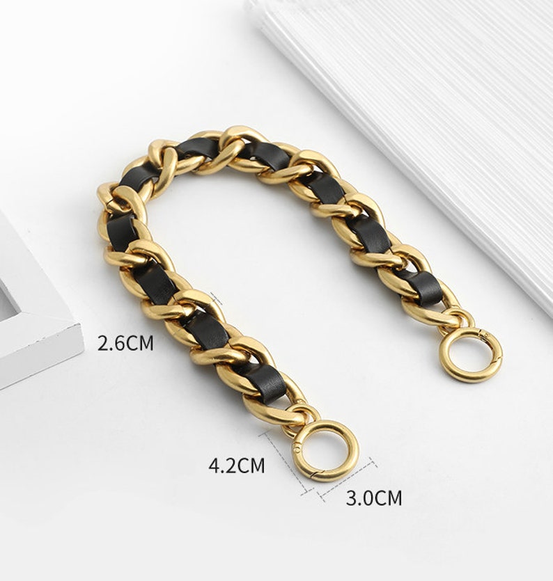 Peut inclure: Une cha&icirc;ne en m&eacute;tal dor&eacute; entrelac&eacute;e d'une bande en similicuir noir. La cha&icirc;ne comporte deux anneaux circulaires &agrave; ressort aux extr&eacute;mit&eacute;s. Les dimensions indiqu&eacute;es sont 2,6 cm, 4,2 cm et 3,0 cm, pr&eacute;cisant la taille des maillons et des anneaux.