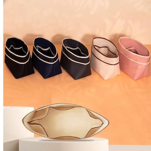 Pode incluir: Uma coleção de cinco organizadores de tecido em várias cores: preto, azul marinho, cinza escuro, bege e rosa. Cada organizador tem uma borda branca contrastante e um design estruturado. Um organizador adicional é exibido aberto, revelando compartimentos internos.