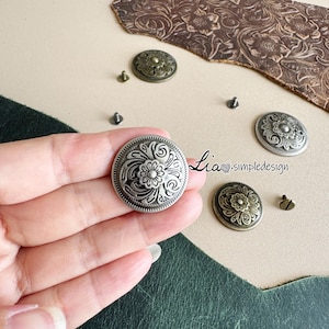 Puede incluir: Primer plano de botones decorativos de metal con diseños florales. Los botones son de varios tonos plateados y bronce, con intrincados patrones en relieve. También se ven pequeños tornillos, lo que sugiere que sirven para fijar los botones. La imagen incluye el texto "Lia simpledesign".