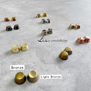 May include: A selection of metal cone-shaped rivets in gold, bronze, silver, and copper hues, arranged on a grey surface. The image features the text "Bronze" and "Light Bronze" near the rivets.