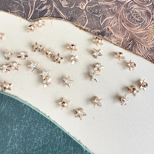 Puede incluir: Una colección de adornos en forma de estrella de metal dorado con diamantes de imitación transparentes en el centro. Las estrellas están dispersas sobre una superficie beige claro.
