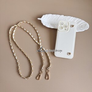 May include: White phone case with a gold chain strap. The case has a textured surface and gold accents. The chain strap is white with gold chain links. The phone case and strap are on a neutral background.