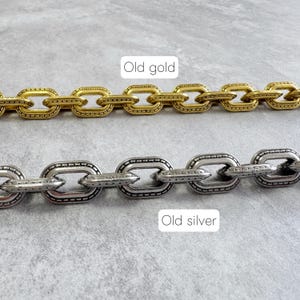 Può includere: Primo piano di due collane a maglie. Una è oro antico, l'altra argento antico. Entrambe hanno un design a maglie ovali con un motivo strutturato. Lo sfondo è una superficie grigio chiaro.