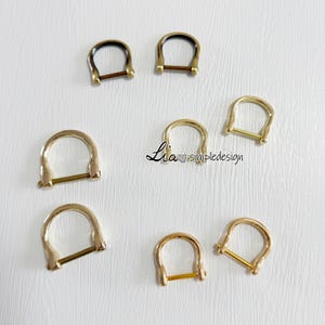 May include: Assortment of gold and black metal D-rings. The D-rings are different sizes and are arranged in a circle on a white surface.