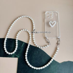 May include: Clear phone case with a white heart design and a pearl beaded lanyard. The lanyard is made of white pearls and silver-toned accents. The text "Lia simpledesign" is printed on the case.
