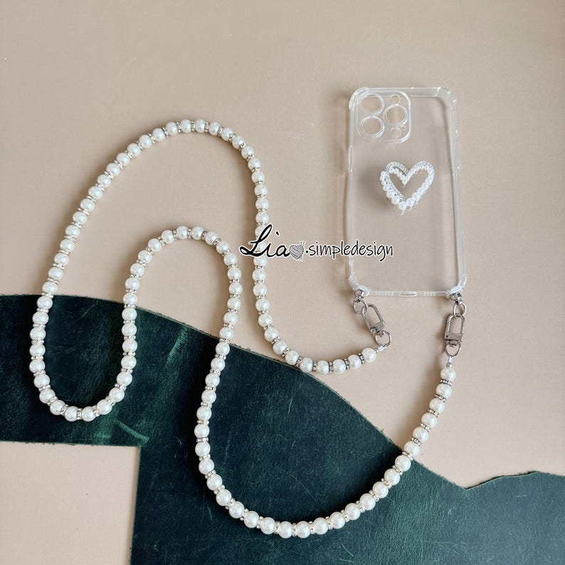 May include: Clear phone case with a white heart design and a pearl beaded lanyard. The lanyard is made of white pearls and silver-toned accents. The text "Lia simpledesign" is printed on the case.