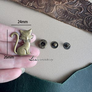 Peut inclure: Un charme de chat en métal doré avec un nœud papillon, mesurant 26 mm sur 24 mm. Le charme est entouré de quatre plus petites pièces métalliques, dont deux boutons-pression ronds et deux pièces décoratives.