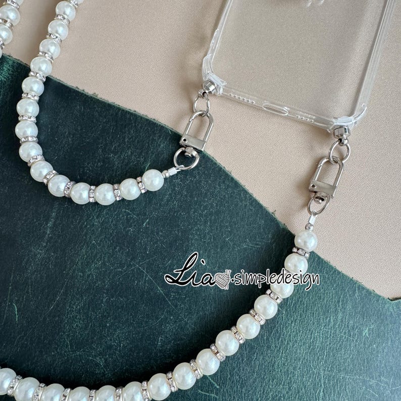 May include: Clear phone case with a pearl and rhinestone beaded strap. The strap is attached to the case with silver-colored metal clasps. The text "Lia simpledesign" is visible on the bottom of the image.