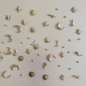 Puede incluir: Un surtido de adornos de temática celestial, que incluyen lunas crecientes, estrellas y rayos de sol, en tonos plateados y dorados. El diseño presenta varios tamaños y estilos de estos elementos decorativos. El texto "Lia simpledesign" es visible.