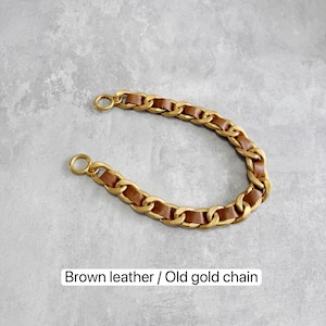 Peut inclure: Une bandouli&egrave;re incurv&eacute;e compos&eacute;e de maillons m&eacute;talliques dor&eacute;s entrelac&eacute;s avec une lani&egrave;re en cuir marron. La sangle comprend un anneau m&eacute;tallique circulaire &agrave; chaque extr&eacute;mit&eacute; pour la fixation. Le texte "Brown leather / Old gold chain" est visible.