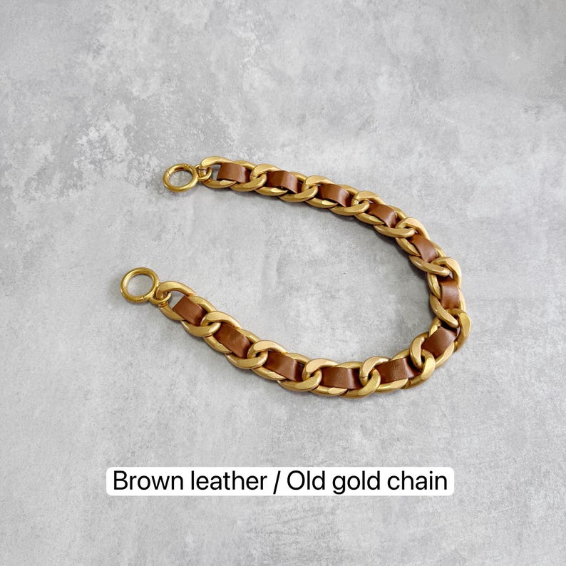 Peut inclure: Une bandouli&egrave;re incurv&eacute;e compos&eacute;e de maillons m&eacute;talliques dor&eacute;s entrelac&eacute;s avec une lani&egrave;re en cuir marron. La sangle comprend un anneau m&eacute;tallique circulaire &agrave; chaque extr&eacute;mit&eacute; pour la fixation. Le texte "Brown leather / Old gold chain" est visible.