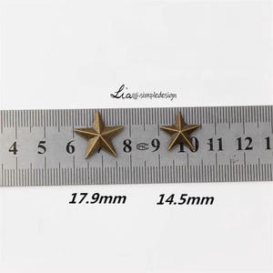 May include: Two gold-toned metal star-shaped embellishments, measured against a ruler. The larger star measures 1.79 inches and the smaller star measures 1.45 inches.