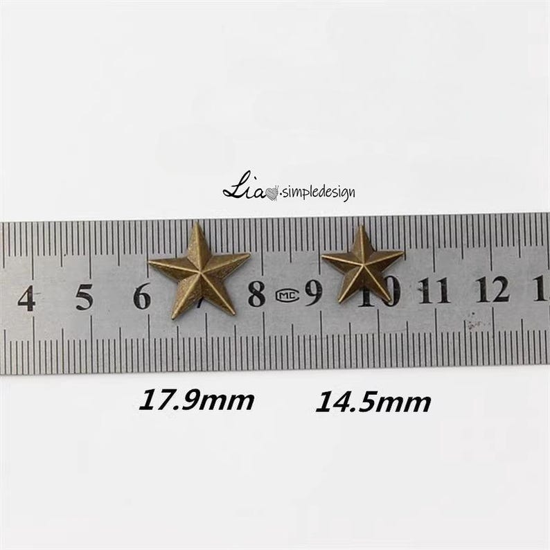 May include: Two gold-toned metal star-shaped embellishments, measured against a ruler. The larger star measures 1.79 inches and the smaller star measures 1.45 inches.