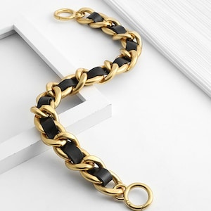 Peut inclure: Un bracelet &agrave; cha&icirc;ne en m&eacute;tal dor&eacute; compos&eacute; de maillons ovales &eacute;pais et entrelac&eacute;s, entrelac&eacute;s d'une bande de cuir noir lisse. Le bracelet comprend un fermoir &agrave; ressort circulaire &agrave; chaque extr&eacute;mit&eacute;, cr&eacute;ant un design flexible et textur&eacute;.