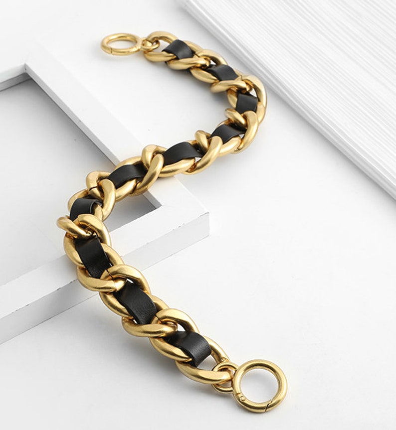 Peut inclure: Un bracelet &agrave; cha&icirc;ne en m&eacute;tal dor&eacute; compos&eacute; de maillons ovales &eacute;pais et entrelac&eacute;s, entrelac&eacute;s d'une bande de cuir noir lisse. Le bracelet comprend un fermoir &agrave; ressort circulaire &agrave; chaque extr&eacute;mit&eacute;, cr&eacute;ant un design flexible et textur&eacute;.