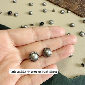 May include: A collection of antique silver mushroom punk rivets featuring a smooth, rounded top and a ribbed edge. The metal fasteners are scattered across a surface, with two rivets resting on a palm. The text reads Antique Silver Mushroom Punk Rivets.
