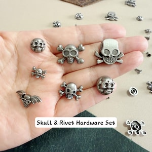 May include: A collection of silver-toned metal rivets and studs featuring various skull designs, including some with crossbones and wings. The set includes circular and shaped hardware pieces. The text Skull & Rivet Hardware Set is visible.