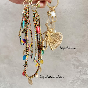 May include: Three bag charms featuring braided multi-coloured cords in red, yellow, and black, adorned with various small metal and plastic charms, including a large gold-toned heart pendant, a white star, and a small red fish-shaped charm.