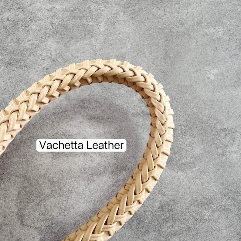 May include: Close-up of a braided Vachetta leather handle, in a light beige hue. The handle is curved, featuring a detailed woven design. The text "Vachetta Leather" is displayed in a white rectangle.