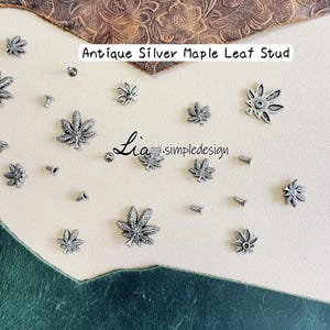 Könnte beinhalten: Antike silberne Ahornblatt-Nieten, verstreut auf einer hellen Oberfläche. Die Nieten sind klein, detailliert und haben ein Vintage-Aussehen. Der Text "Antique Silver Maple Leaf Stud" ist sichtbar.