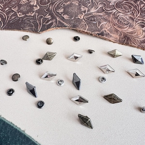 Può includere: Assortimento di borchie metalliche a forma di diamante di colore argento, oro e bronzo. Le borchie sono sparse su una superficie bianca.
