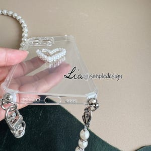 May include: Clear phone case with a pearl heart design and a pearl strap. The case has silver hardware and the text "Lia simpledesign". The pearl strap is attached to the case with silver clasps.