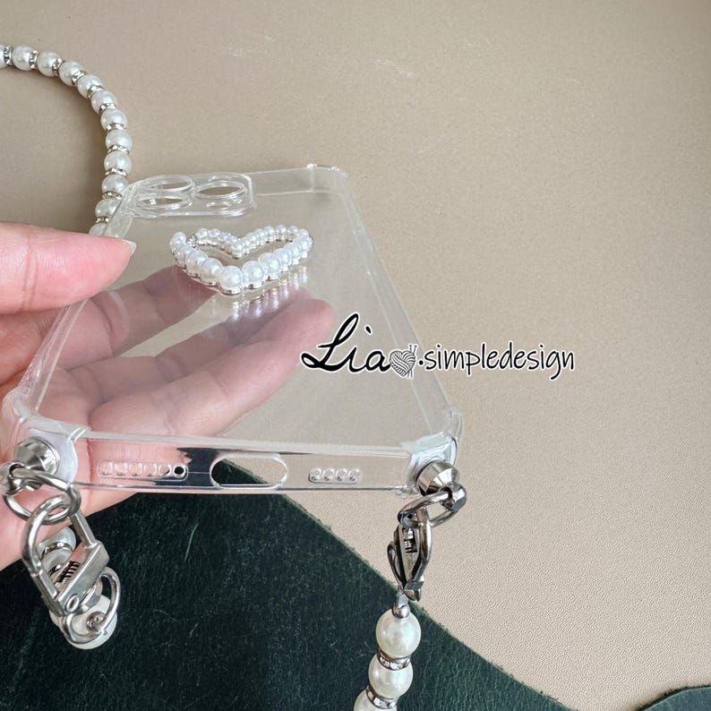 May include: Clear phone case with a pearl heart design and a pearl strap. The case has silver hardware and the text "Lia simpledesign". The pearl strap is attached to the case with silver clasps.