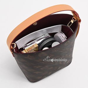 Puede incluir: Un bolso de cubo de patrón marrón y negro con una correa de cuero marrón claro y herrajes dorados. El bolso está abierto y tiene varios artículos en su interior, incluyendo un recipiente de plástico transparente, un recipiente redondo negro y un recipiente blanco pequeño.