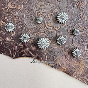 Può includere: Primo piano di abbellimenti metallici argentati a forma di fiore sparsi su una superficie in pelle marrone con goffratura floreale. La pelle ha un aspetto invecchiato. L'angolo in basso a sinistra mostra una superficie bianca con il testo "Lia simpledesign".