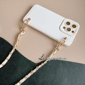 May include: White phone case with a gold chain strap. The case has a textured surface and a camera cutout. The chain is made of gold links intertwined with white leather. The text "Lia simpledesign" is visible.