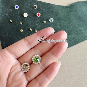 Peut inclure: Gros plan sur divers petits ornements décoratifs en strass. Les ornements présentent une pierre centrale colorée entourée de petits strass transparents. Les couleurs incluent le bleu, le rouge, le vert et le transparent. Le texte "Lia simpledesign" est également présent.