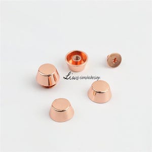 May include: Rose gold metal cone-shaped furniture feet and a screw. The feet have a flat top and a flared base. The screw has a cross-shaped indentation. The text "Lia. simpledesign" is visible.