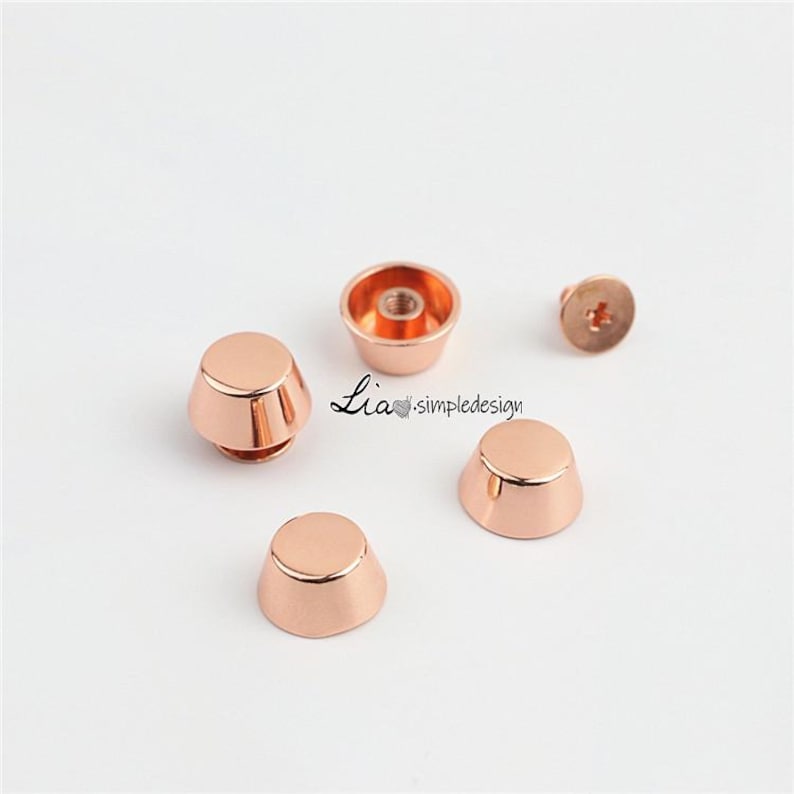 May include: Rose gold metal cone-shaped furniture feet and a screw. The feet have a flat top and a flared base. The screw has a cross-shaped indentation. The text "Lia. simpledesign" is visible.