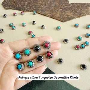 May include: A collection of antique silver decorative rivets featuring a flower-shaped metal base with a central cabochon stone in turquoise, red, or black. The rivets are scattered across a surface, with several held in a palm.