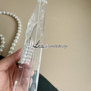 May include: Clear phone case with the text "Lia simpledesign" in black script. The case has a pearl beaded strap attached. The phone case is made of clear plastic and has a raised design on the side.