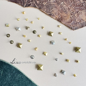 May include: A collection of metal studs in diverse shapes and sizes, including pyramid and round designs. The studs are in gold, silver, and bronze tones, scattered on a neutral surface. The image includes the text "Lia simpledesign".