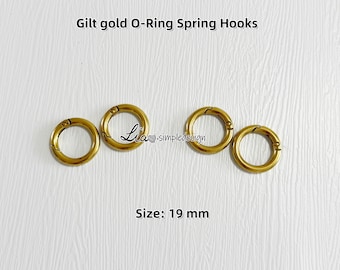 Puede incluir: Cuatro ganchos de resorte dorados de 19 mm de diámetro, sobre un fondo blanco. El texto "Gilt gold O-Ring Spring Hooks" y "Size: 19 mm" son visibles. Los ganchos son circulares con un cierre de resorte.