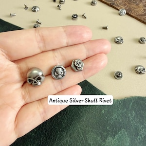 May include: Three antique silver-toned metal rivets featuring skull designs, including one rounded skull, one circular skull, and one hexagonal skull. The text Antique Silver Skull Rivet is visible. These small hardware fasteners are metallic grey.