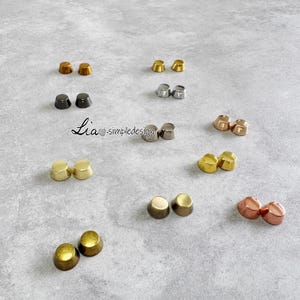 May include: A collection of metal stud earrings in different colours, such as gold, silver, bronze, and black. The earrings are displayed on a grey surface. The image includes the text "Lia simpledesign".