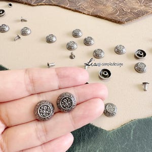 May include: Close-up of antique silver-colored metal rivets with a decorative circular design. The rivets are scattered on a light beige surface, with some held in a hand. The image includes the text "Lia simpledesign".